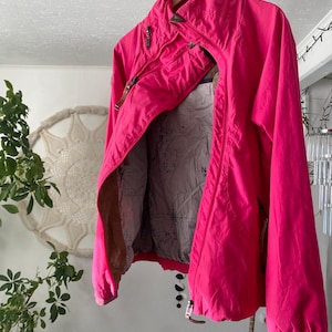 Può includere: Giacca rosa acceso con collo alto e chiusura a zip. La giacca è appesa a una gruccia di legno con un motivo a zampa. La fodera interna presenta un disegno a mappa.