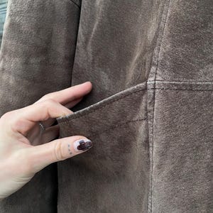 May include: Close-up of a brown suede jacket. The jacket features a pocket and visible stitching details. The suede has a soft, textured appearance. The jacket appears to be well-worn.