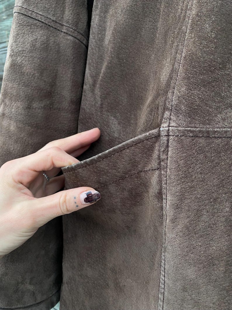 May include: Close-up of a brown suede jacket. The jacket features a pocket and visible stitching details. The suede has a soft, textured appearance. The jacket appears to be well-worn.