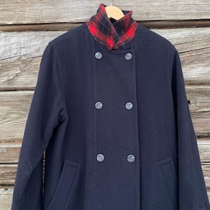 May include: A navy blue double-breasted coat with a red and black plaid collar. The coat has four dark buttons and two front pockets. The coat is hanging on a wooden hanger against a weathered wood background.
