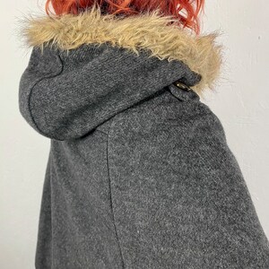 May include: A gray hooded cape with a faux fur trim around the hood. The cape has a toggle closure at the neck. The fabric appears to be a wool blend. The cape is a stylish outerwear garment.