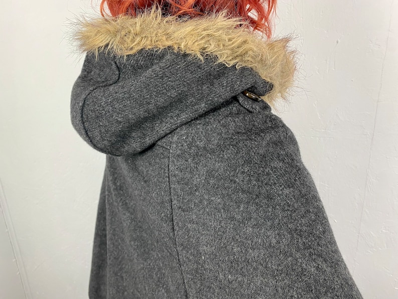 May include: A gray hooded cape with a faux fur trim around the hood. The cape has a toggle closure at the neck. The fabric appears to be a wool blend. The cape is a stylish outerwear garment.