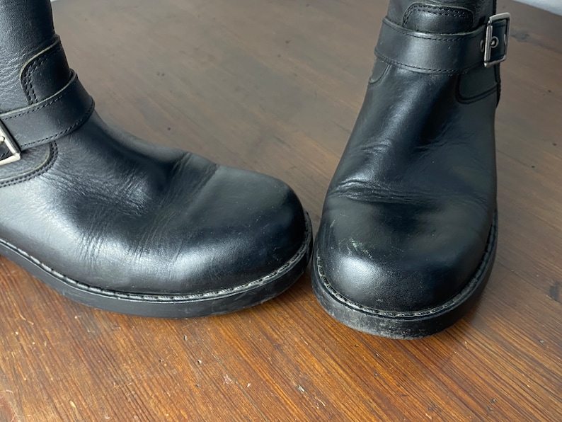 May include: A pair of black leather ankle boots with a buckle strap. The boots have a slightly worn look and are made of a smooth leather.