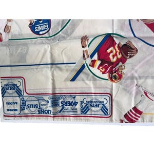 Puede incluir: Una tela blanca con un diseño colorido que presenta logotipos y jugadores de la NFL. Los logotipos incluyen los Jets, los Buccaneers, los Packers, los 49ers, los Eagles, los Oilers, los Lions, los Seahawks, los Colts, los Bills, los Browns, los Rams, los Raiders y los Giants.