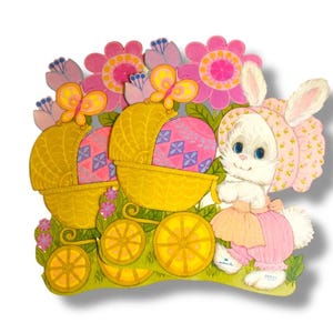 May include: A vintage Easter-themed die-cut decoration featuring a white bunny in a pink bonnet and dress, pushing a yellow carriage with Easter eggs. The background includes pink flowers and butterflies, creating a cheerful spring scene.