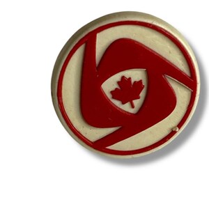 May include: A round, cream-colored pin with a red design. The design features a stylized swirl with a red maple leaf in the center. The pin has a red border.