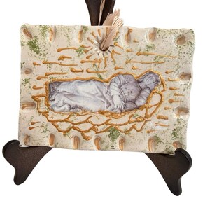 May include: A rectangular ceramic wall hanging with a depiction of a reclining figure. The artwork features a cream-colored background with gold and green accents, and is mounted on a dark brown wooden stand.