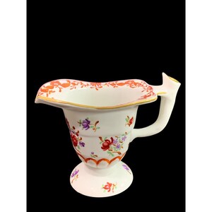 May include: A white porcelain creamer with a scalloped rim and a floral design in shades of red, orange, purple, and green. The creamer has a handle and a base with a scalloped edge.
