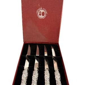 May include: A set of four silver-toned knives with textured handles featuring a grape vine motif, presented in a deep red box. The interior lid of the box displays the white text Arthur Court Designs California with a circular logo containing the number 60.