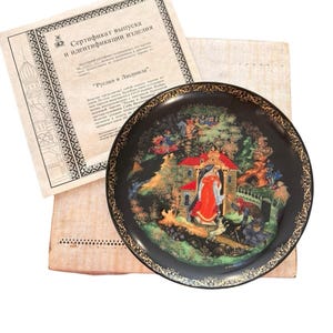 May include: A decorative black plate with a detailed, colorful scene featuring figures in a landscape, possibly a fairy tale. The plate has a gold rim and is accompanied by a certificate with Russian text.