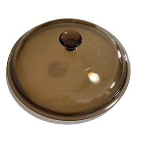 Puede incluir: Una tapa redonda de vidrio marrón con un pequeño pomo marrón oscuro. La tapa tiene un borde de metal plateado. El vidrio transparente permite ver el contenido. Utensilios de cocina.