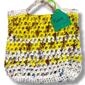 May include: A tote bag crocheted from strips of white and yellow plastic bags, featuring an open-weave pattern. A green tag attached with a lime green string displays the handwritten text Plarn (Plastic Yarn) in black ink on the front surface.