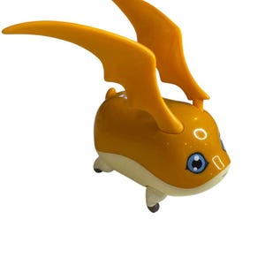 May include: A yellow and cream-colored toy figure with large, wing-like structures on its back. The figure has blue eyes and small, rounded feet. The toy is a cartoon-like character.