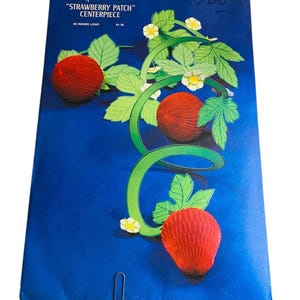 May include: A Hallmark "Strawberry Patch" centerpiece on a blue background. The design features three red paper strawberries, green vines, and white flowers. The centerpiece is 30 inches long.