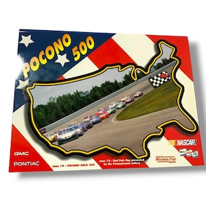 May include: A rectangular Pocono 500 promotional sign featuring a red, white, and blue background with a central United States map outline containing a photograph of race cars on a track. Text includes GMC, Pontiac, and June 19 - Pocono ARCA 200.