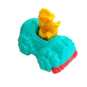 May include: A small plastic toy vehicle in a bright teal color with a textured, rock-like surface. A yellow dinosaur-like figure is seated in the open cockpit of the vehicle. The toy features a red bumper at the front and a red axle at the rear.