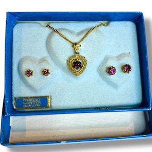 May include: A jewelry set in a blue box, featuring a gold-toned necklace with a heart-shaped pendant and matching earrings. The pendant and earrings have a cluster of purple gemstones. The box has a blue label with text.