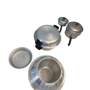 May include: A collection of vintage aluminum cookware, including a large pot with black handles, two smaller pots with lids, a shallow dish, and a conical-shaped container. The items are arranged on a white background.