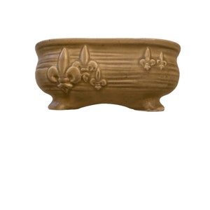 May include: A light brown ceramic planter with a decorative design of fleur-de-lis. The planter has a rounded oval shape and three feet.