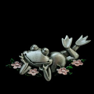 May include: A silver-colored frog figurine, reclining among pink flowers with green leaves. The frog has a relaxed expression and is lying on its back. The figurine is likely a decorative item.