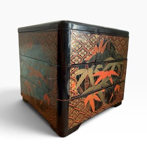 May include: A three-tiered square lacquer box with a glossy black finish. The exterior features intricate gold and orange painted designs of bamboo stalks and foliage set against a repeating geometric diamond pattern on each drawer front.