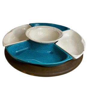May include: A serving dish with five sections, including four white and one turquoise blue, arranged around a central bowl. The sections have textured surfaces and are set on a round, brown base, ideal for snacks or appetizers.