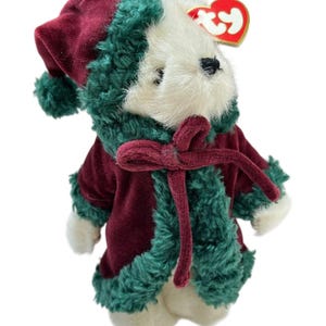 May include: A plush teddy bear wearing a burgundy velvet coat and hat trimmed with green faux fur. The bear has a cream-colored face and paws, and a burgundy bow. A red heart-shaped tag is attached to the bear's ear.
