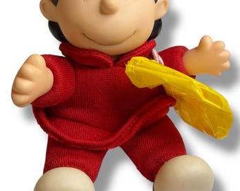 May include: A Lucy doll from the Peanuts comic strip. The doll is wearing a red outfit and has black hair. She is holding a yellow object. The doll's feet are white with black accents.