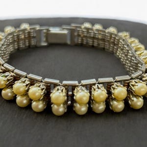 May include: A vintage bracelet featuring a silver-tone metal mesh band, adorned with creamy-yellow faux pearls. The pearls are arranged in clusters of three. A classic piece of jewellery.