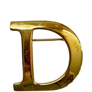 May include: A gold-coloured metal brooch in the shape of the letter "D". The brooch has a pin on the back for attachment. The surface shows signs of wear, adding to its vintage appeal. The brooch is a classic accessory.