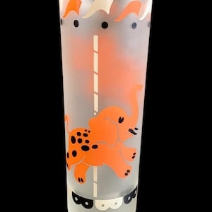 May include: A frosted glass with an orange elephant on a carousel. The elephant has black spots and is riding on a white pole with black and white stripes. The glass has a black and white design around the top and bottom.