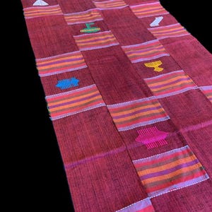 May include: A long, rectangular, woven textile in shades of red, with colorful horizontal stripes and embroidered geometric shapes. The stripes include orange, purple, and pink. The textile is likely a runner or a decorative rug.
