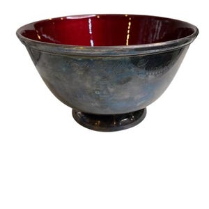 Puede incluir: Un cuenco plateado con interior rojo intenso. El cuenco tiene un borde ancho y una base redondeada. El exterior tiene texto grabado. Una pieza decorativa para servir o exhibir.