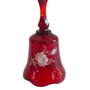 May include: A ruby red glass bell with a long handle. The bell features a white floral design and vertical ribbed detailing. The bell is a decorative item, likely for display or collecting.