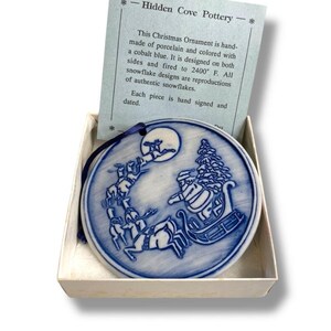 May include: A cobalt blue porcelain Christmas ornament featuring Santa Claus in a sleigh pulled by reindeer, with a full moon in the background. The ornament is in a box with a card that reads "Hidden Cove Pottery".