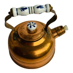 May include: A copper tea kettle with a white and blue floral patterned handle and lid knob. The kettle has a round body and a spout with a white whistle. The handle is made of metal and is attached to the kettle.
