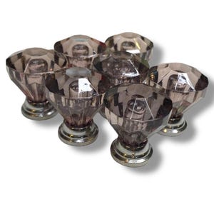 Puede incluir: Un conjunto de siete pomos de armario de cristal facetado de color amatista. Cada pomo tiene forma de diamante y se asienta sobre una base de metal plateado. Los pomos están dispuestos muy juntos, mostrando sus superficies reflectantes.
