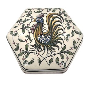 May include: Hexagonal ceramic trinket box with a rooster design. The rooster is yellow, green, blue, and red with a white background. The box is decorated with green vines and red dots.