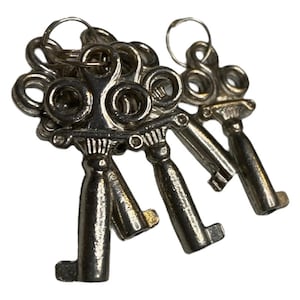 May include: A collection of antique silver-colored keys on a white background. The keys have ornate, decorative tops and are attached to small metal rings. The key handles have a classic, vintage design.