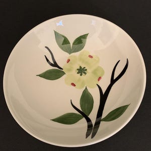 May include: Cream ceramic bowl with a hand-painted floral design. The bowl features a large, pale yellow flower with red accents, green leaves, and black stems. The design is centered on the bowl's interior, creating a decorative and functional piece.