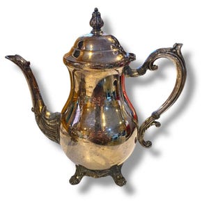 Puede incluir: Una tetera plateada con un pico curvo y un asa ornamentada. La tetera tiene un cuerpo bulboso y se apoya en tres pequeños pies. La tapa está rematada con un adorno decorativo. La tetera es probablemente vintage o antigua.