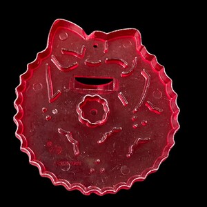 May include: Red plastic Christmas wreath cookie cutter with a bow at the top and a small flower in the center. The cutter has a hole at the top for hanging.