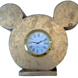 May include: A gold-colored Mickey Mouse-shaped quartz clock. The clock face has Roman numerals and gold-colored hands. The clock is made of a textured material and has a rectangular base.