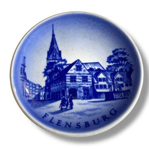 Puede incluir: Pequeño plato decorativo redondo azul y blanco con una ilustración detallada de un edificio con un alto campanario y la palabra "FLENSBURG" en una fuente curva. El plato tiene un borde azul.