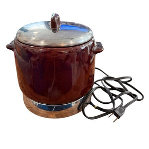 Puede incluir: Una olla de cocción lenta de cerámica de color marrón oscuro con una tapa metálica brillante y dos asas laterales. El aparato cuenta con una base circular y un cable de alimentación negro con un enchufe estándar que sale del lateral de la unidad.