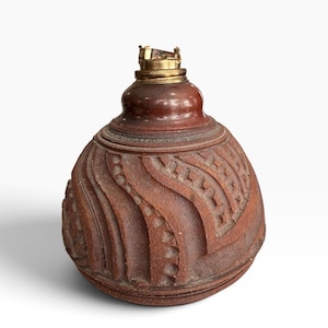May include: A round, reddish-brown ceramic oil lamp base featuring a textured surface with carved, swirling wave patterns and geometric indentations. The top of the vessel is fitted with a metallic brass-colored collar and a small burner mechanism.
