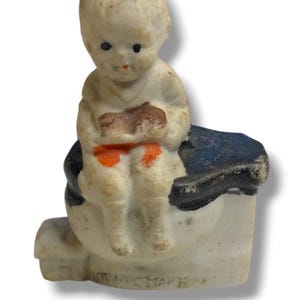 Puede incluir: Una pequeña figura de cerámica de color blanco hueso que representa a un niño sentado en un inodoro. El niño tiene ojos negros pintados y boca roja, vistiendo un conjunto blanco con detalles naranjas. La base del inodoro es blanca con asiento azul oscuro.