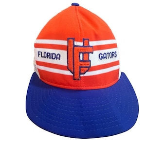 Puede incluir: Una gorra de béisbol roja y azul con un patrón de rayas blancas. La gorra tiene un logotipo bordado en blanco y azul con el texto "FLORIDA" y "GATORS".