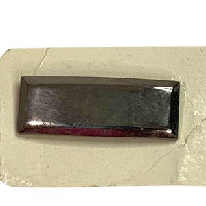 Puede incluir: Un objeto rectangular gris oscuro con un acabado brillante. El objeto está sobre un fondo beige claro. La forma es un rectángulo simple, con un borde ligeramente redondeado.
