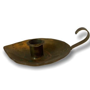 Puede incluir: Un candelero de latón envejecido con forma de hoja. Tiene una copa central para la vela y un asa curvada. El latón presenta un aspecto envejecido, lo que le da un aire vintage. Mide aproximadamente 15 cm de largo.